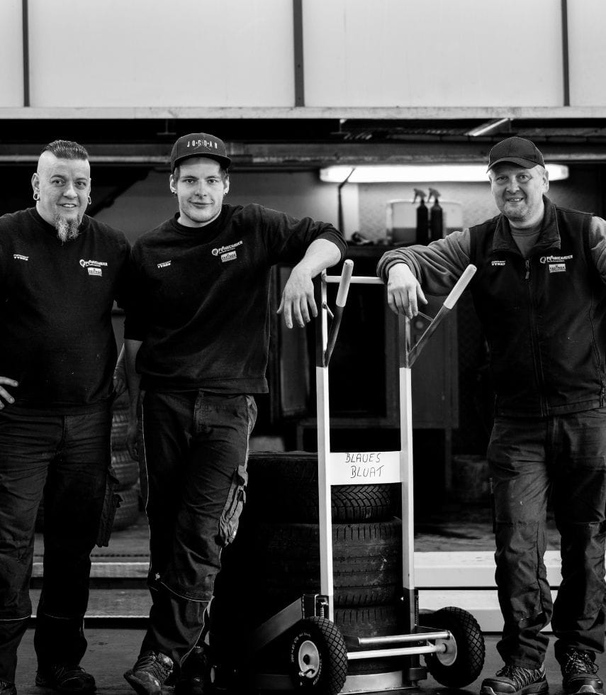 Drei Mitarbeiter stehen gemeinsam vor der Werkstatt und lehnen sich lässig an einen Wagen mit gestapelten Reifen.