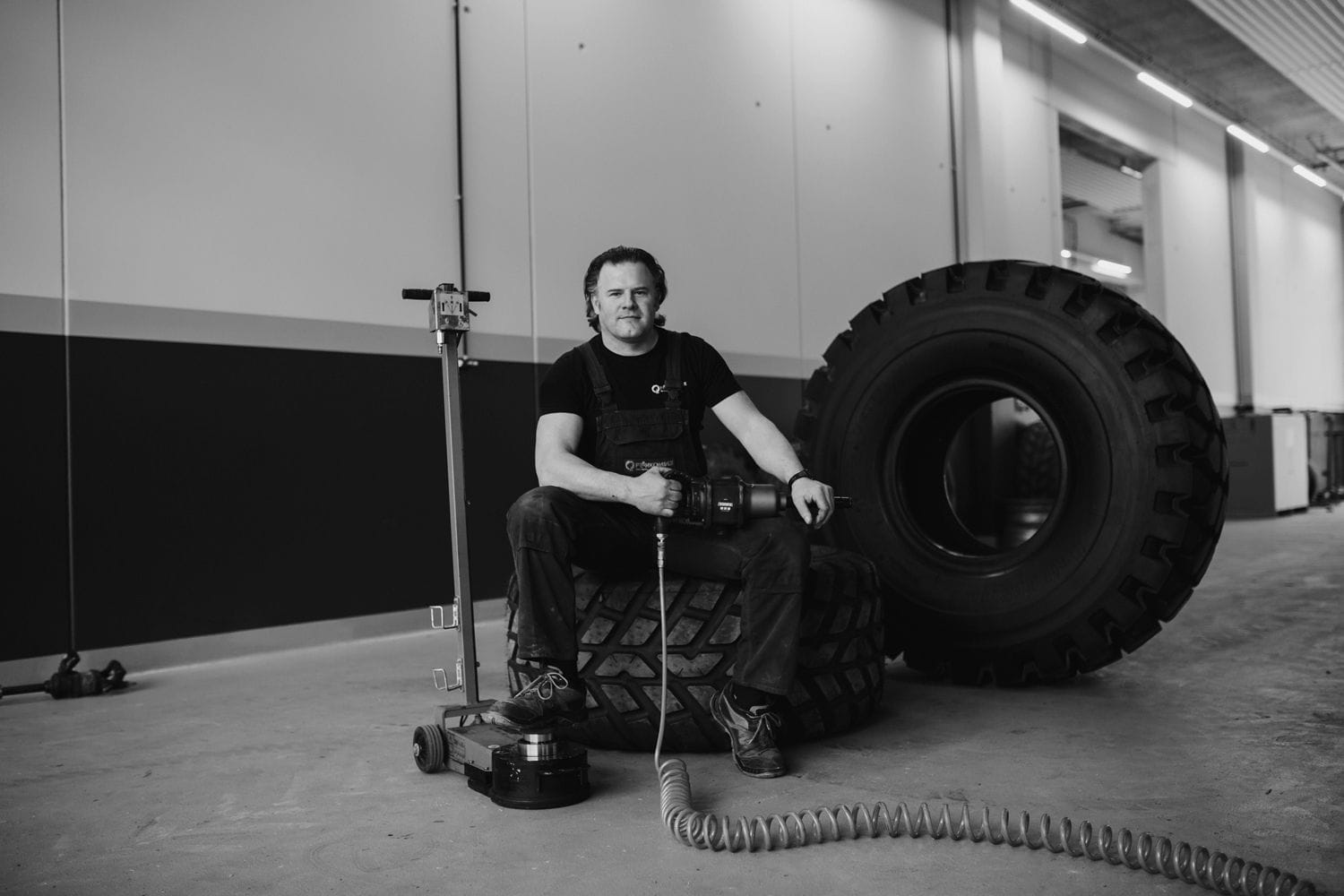 Ein Reifenmonteur sitzt auf einem großen Industriereifen und hält ein Druckluftwerkzeug in der Hand, im Hintergrund weitere Reifen in einer Werkstatthalle.