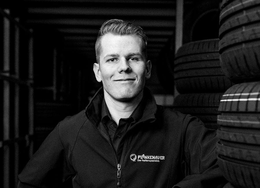 Ein freundlicher Mitarbeiter in Arbeitskleidung steht in einer Werkstatt und blickt direkt in die Kamera.